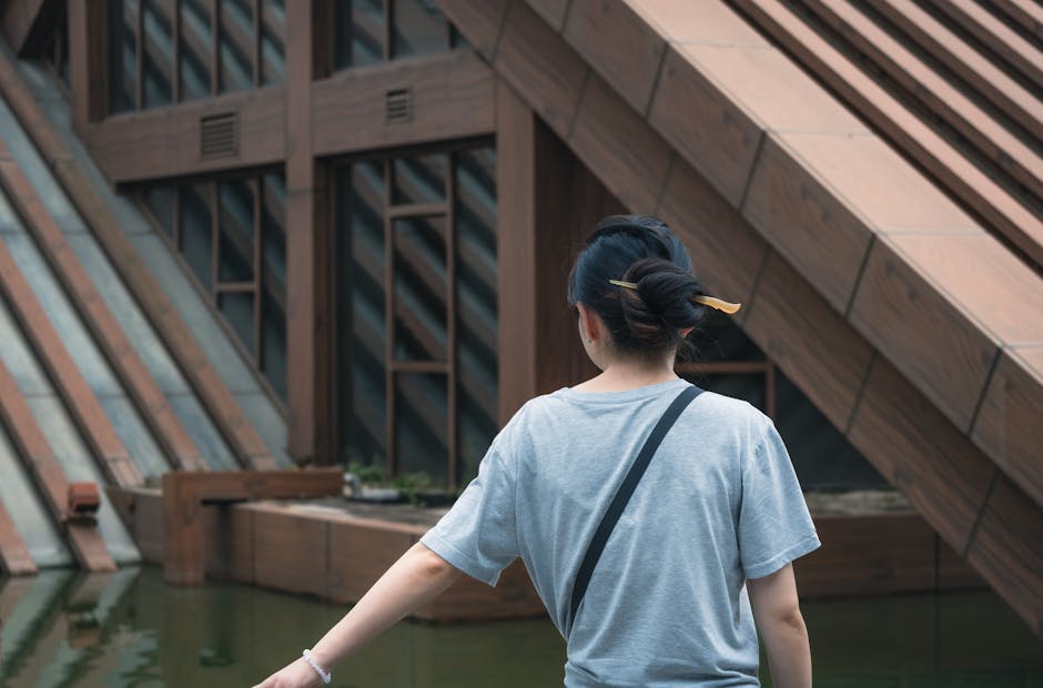 A woman in a gray shirt stands by a modern building with geometric design elements.